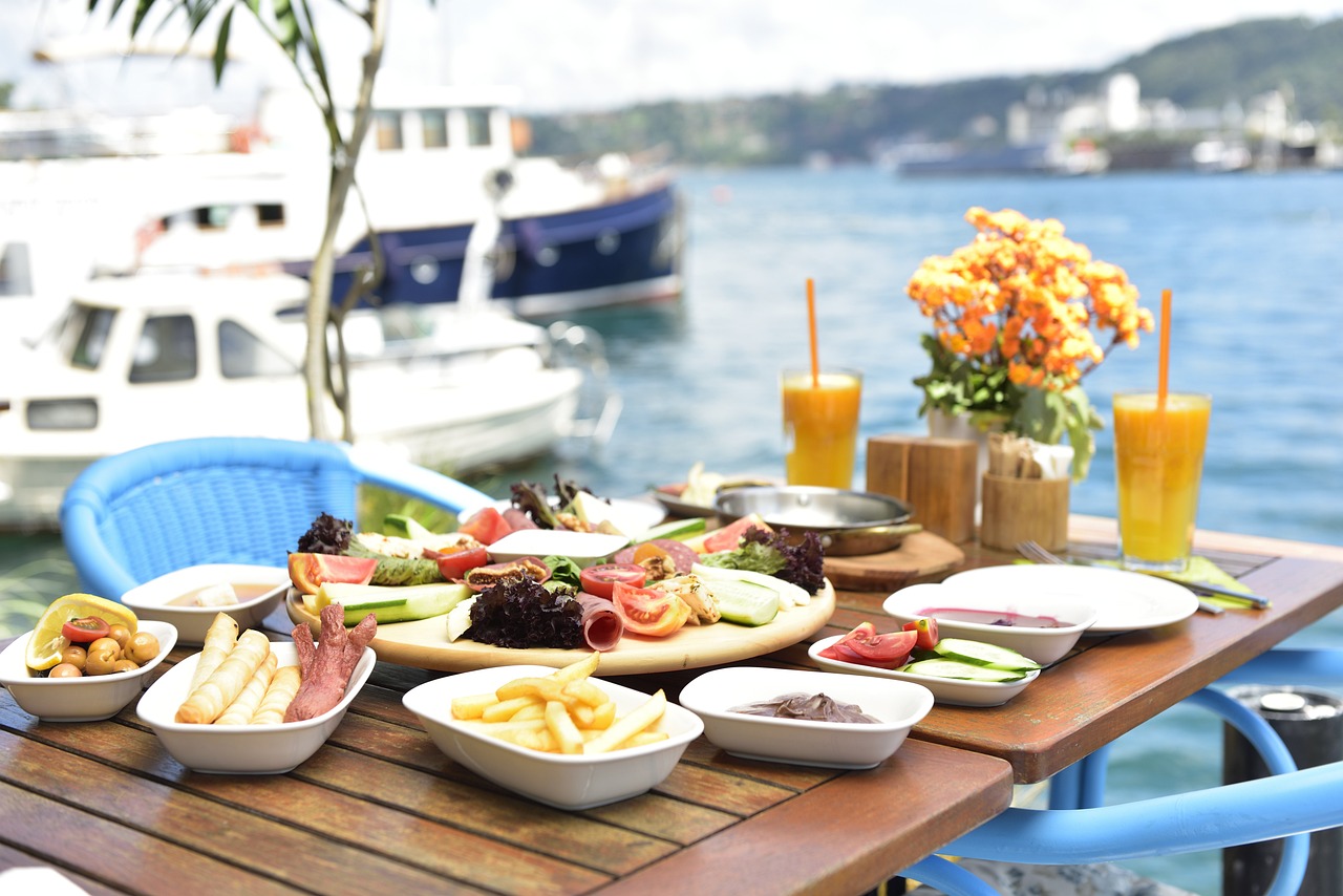 Table de petit-déjeuner turc richement garni (simit, frites, charcuterie, légumes) sur une terrasse en bois avec vue sur le Bosphore et des bateaux amarrés, évoquant l'art de vivre.