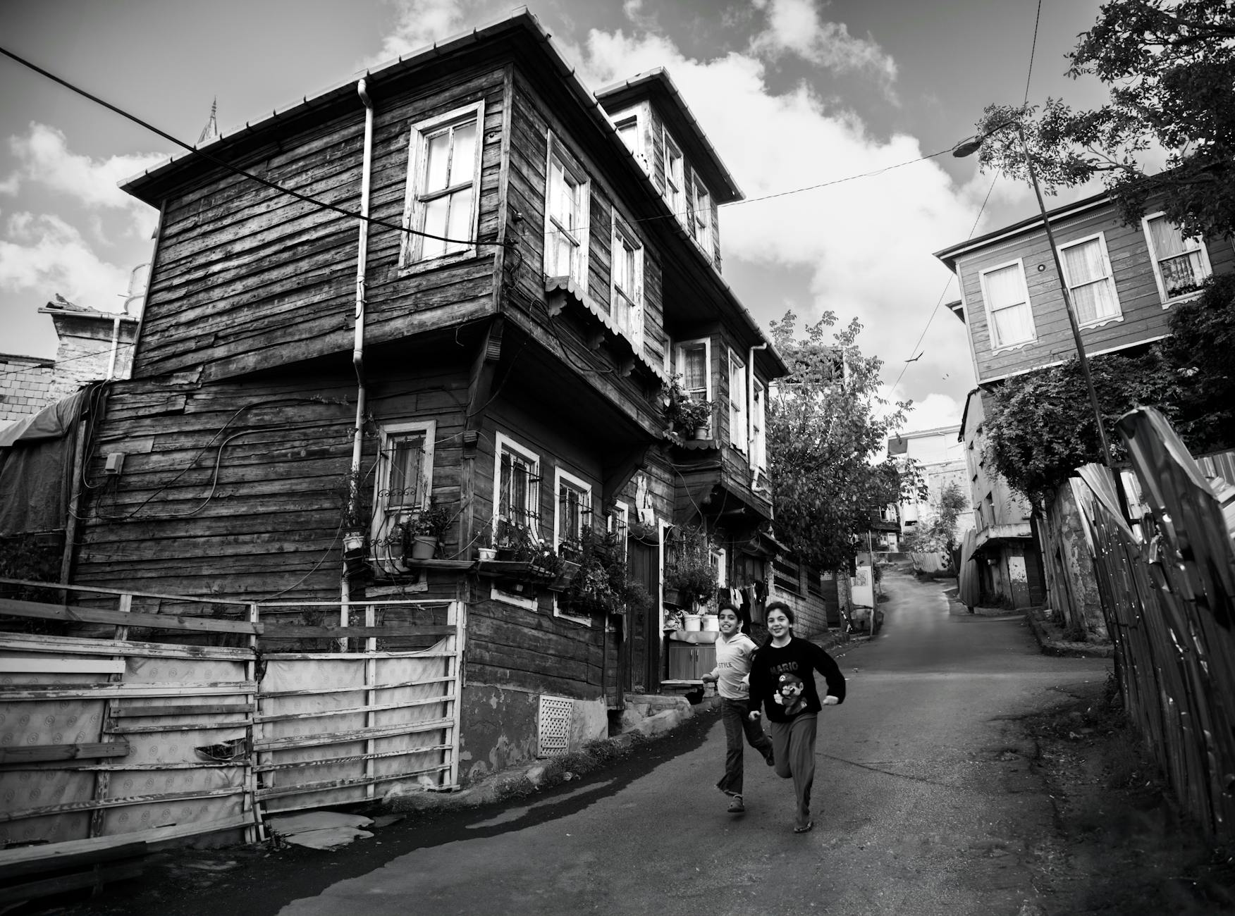 Photographie en noir et blanc d'une rue pavée en pente dans le vieux Beyoğlu à Istanbul, avec une maison en bois traditionnelle en plan rapproché et deux enfants souriants courant vers l'avant.